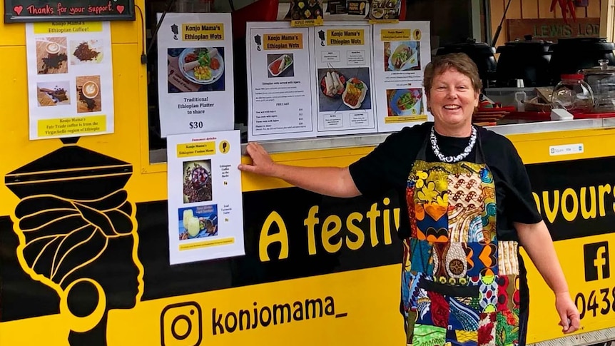 A woman wearing a brightly-coloured apron standing in front of a black and yellow food truck.