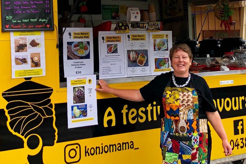 A woman wearing a brightly-coloured apron standing in front of a black and yellow food truck.