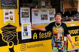 A woman wearing a brightly-coloured apron standing in front of a black and yellow food truck.