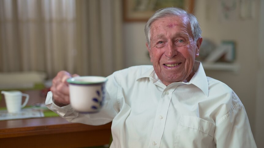 A man in a white shirt smiles and holds up a cup of tea toward the camera