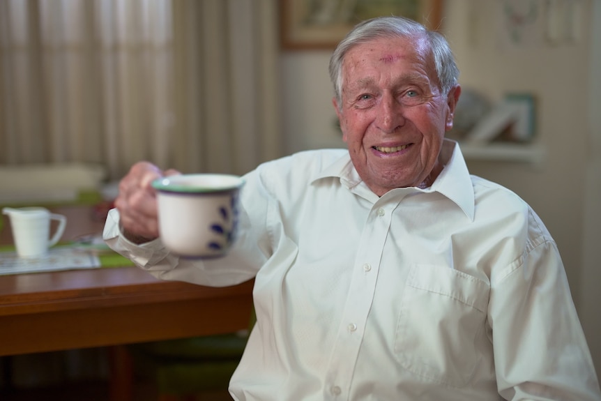 A man in a white shirt smiles and holds up a cup of tea toward the camera