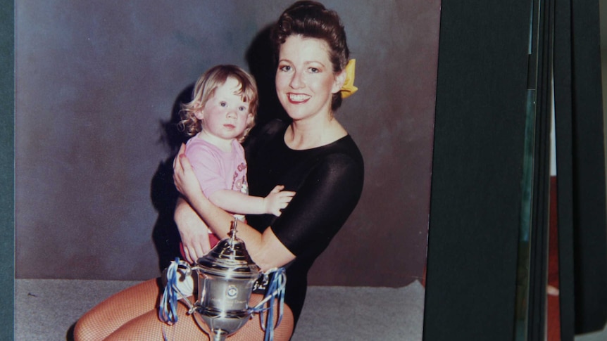 A woman in a black leotard holds her infant daughter in front of a trophy.