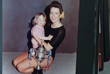 A woman in a black leotard holds her infant daughter in front of a trophy.