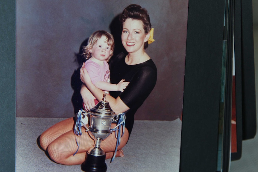 A woman in a black leotard holds her infant daughter in front of a trophy.