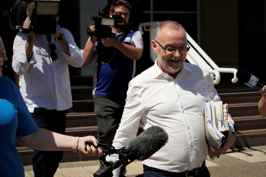 John Lawrence is wearing a white shirt and smiling. There are cameras around him.