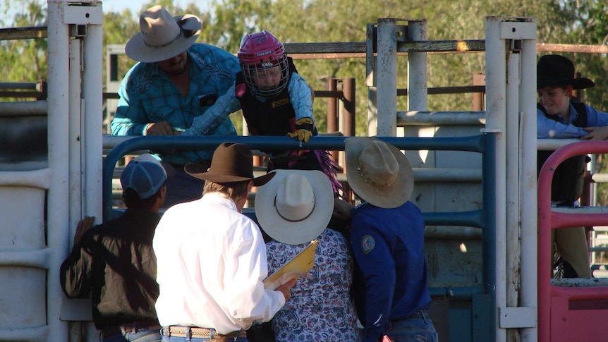 A rider, wearing a full-face helmet, lowers himself onto a poddy calf in a rodeo chute.