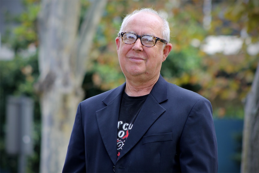 A balding man with glasses stands outside near trees wearing a black coat.