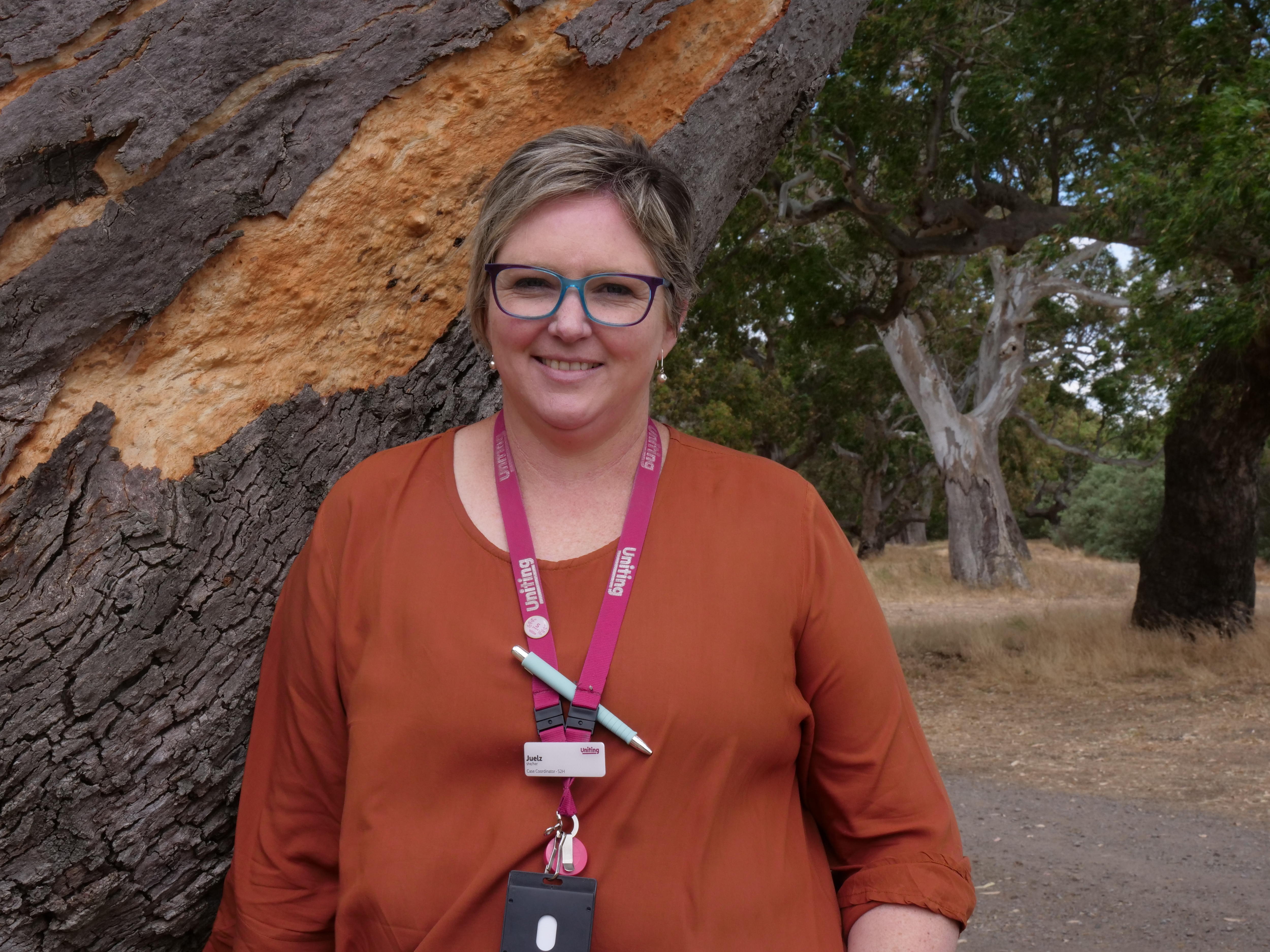 Mujer con camisa naranja de manga larga y gafas. Ella sonríe frente a un árbol. 