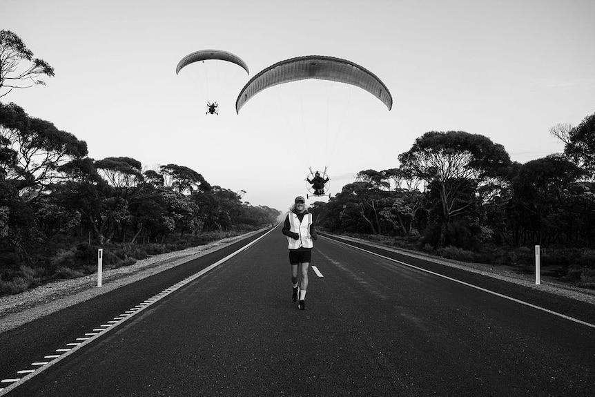 A black and white photo of a man running along the Nullarbor while two men paramotor in the background.