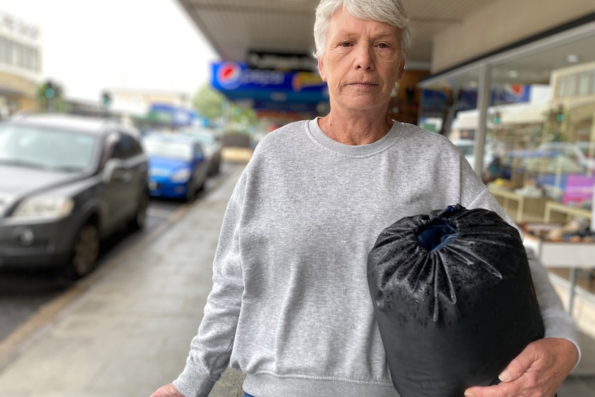 A woman wearing a grey jumper holding a sleeping bag on a street. 