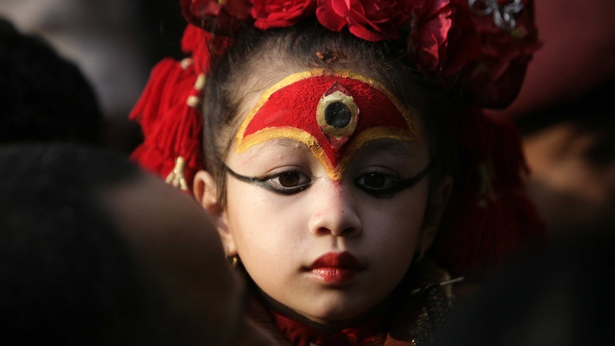 A young girl with a painted forehead and floral headdress looks solemn