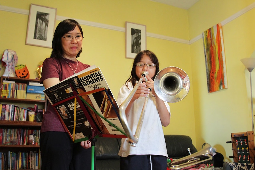 Майка гледа как дъщеря си свири на тромбон в хола на къщата.