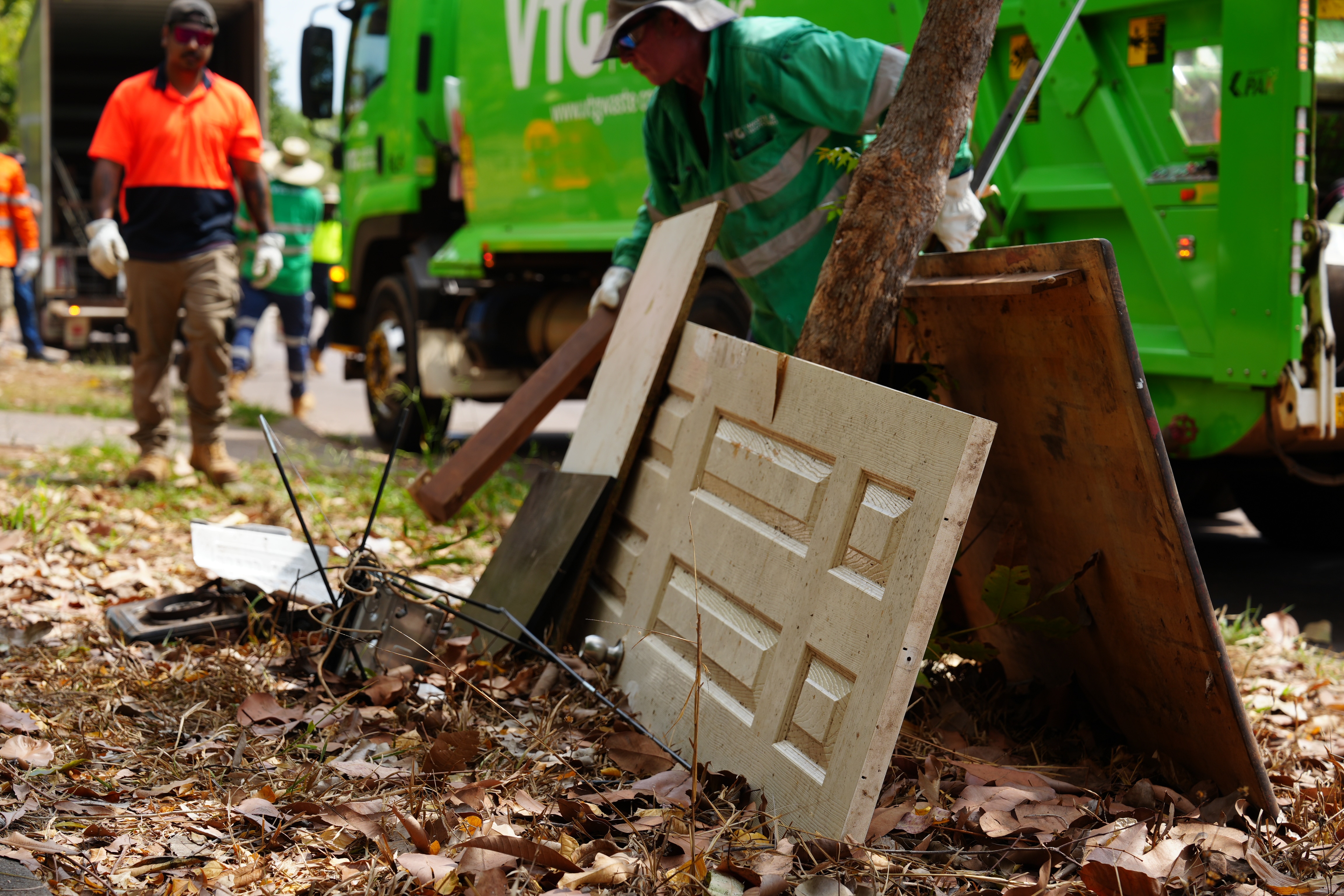 Wooden doors on the curb leaning against a tree, with a green garbage truck on the curb and people in high vis walking toward it