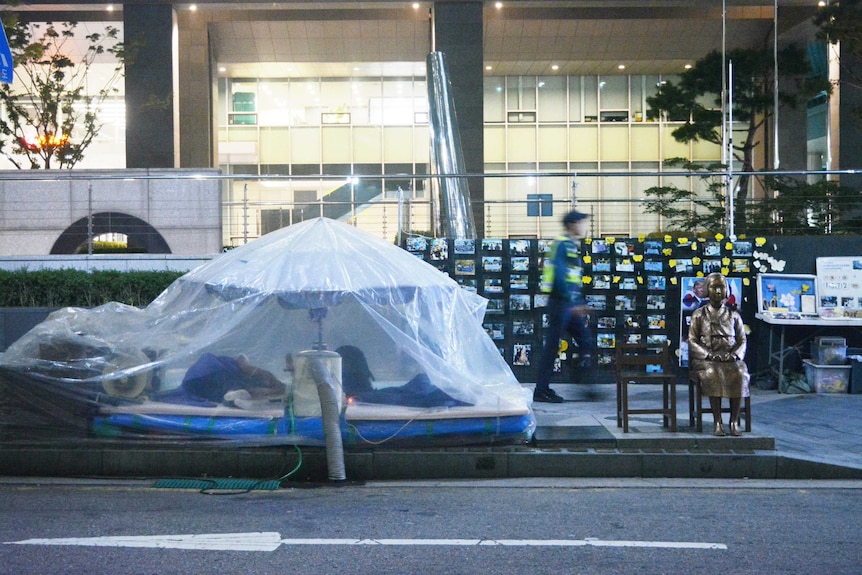 A transparent plastic tent next to a statue as a blurred policeman walks past a large building.