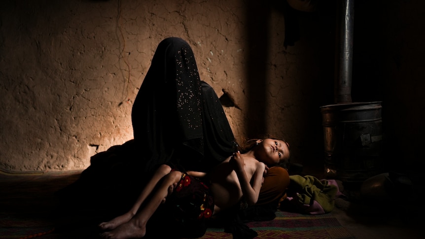 A woman in a niqab sits on the floor holding a wan looking four-year-old girl. 