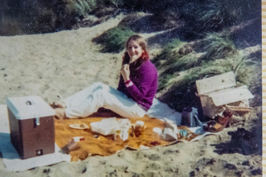 A photo in an album shows a yound woman smiling while sitting on a rug at the beach with an esky