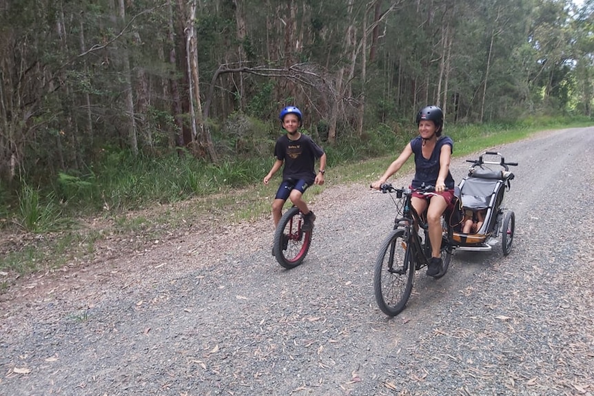 A woman rides a bike towing a child in a cart behind, with a boy riding a unicycle next to her, along a bush track.