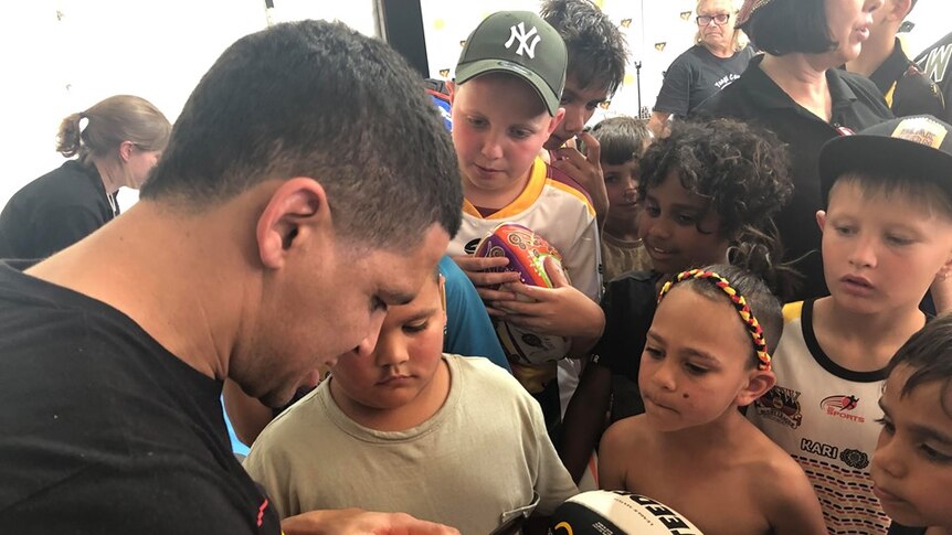 A man signs a football for young fans, with a group of Indigenous children crowded around him