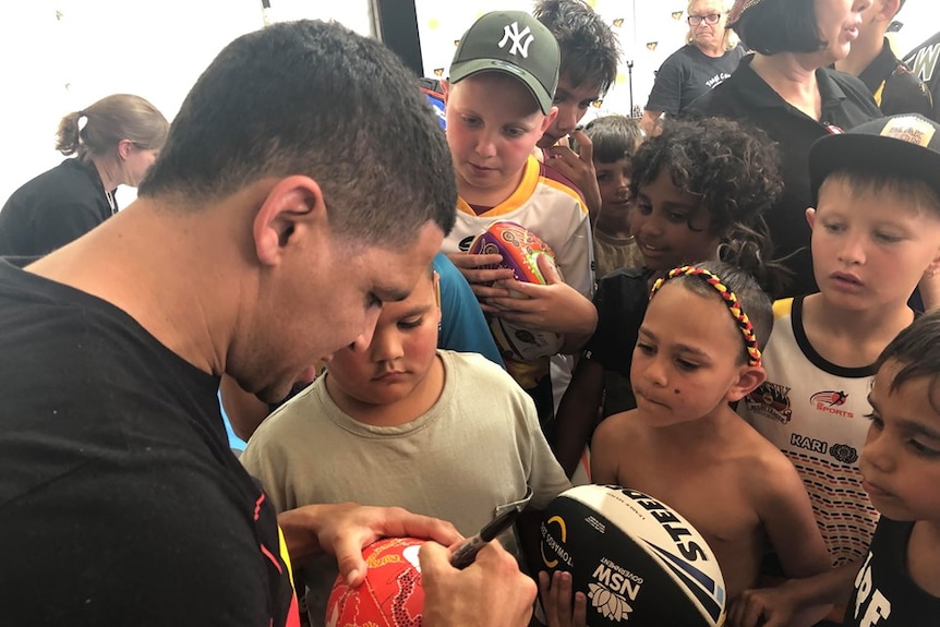 A man signs a football for young fans, with a group of Indigenous children crowded around him