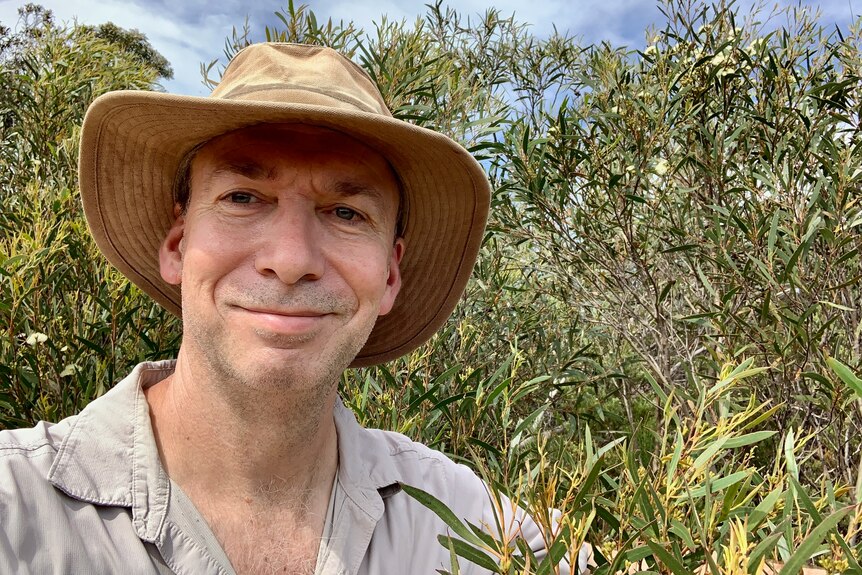 A Caucasian man in a wide brimmed beige hat, James Cook, smiles with his lips closed in front of native trees.