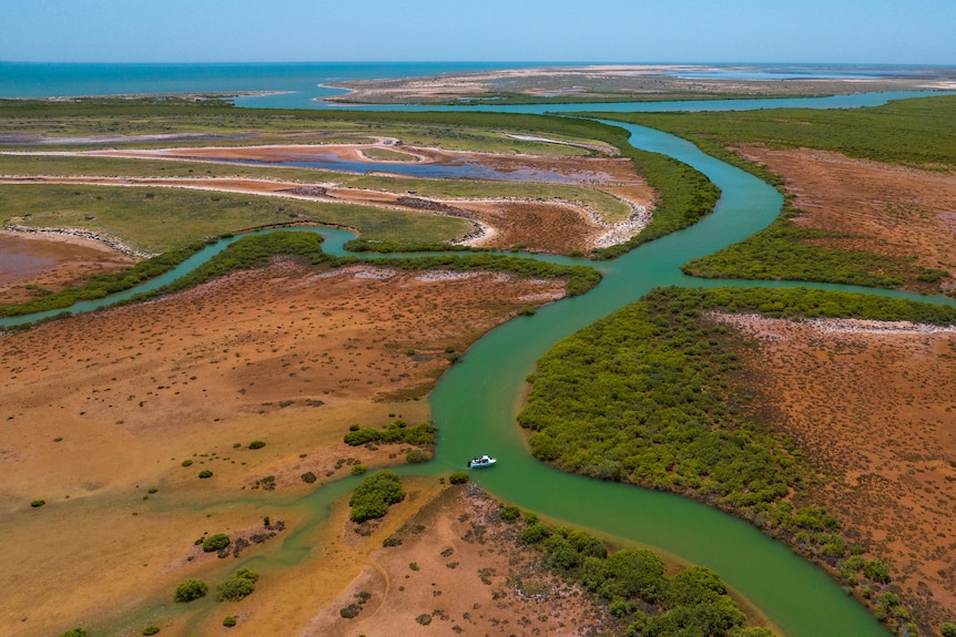 A medium-sized white recreational boat is seen from an aerial view in a green creek cutting through orange land.