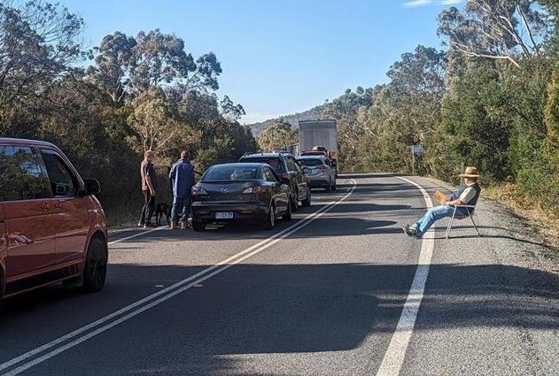 A member of the public sits on a foldup chair on the side of the road waiting for the road to reopen.