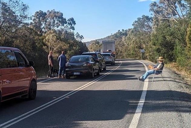 A member of the public sits on a foldup chair on the side of the road waiting for the road to reopen.