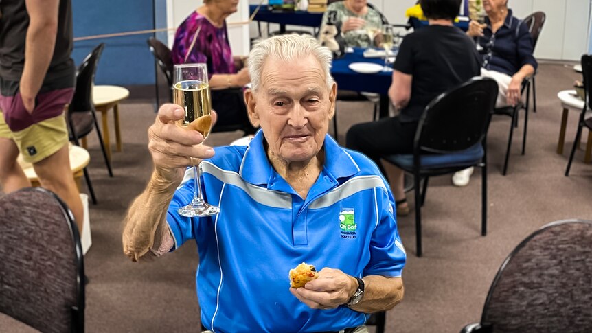 An elderly man in a blue shirt smiles and raises a glass of champagne.