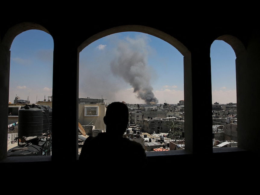 A Palestinian man stands looking through a opening in a building, watching as smoke rises after Israeli strikes.