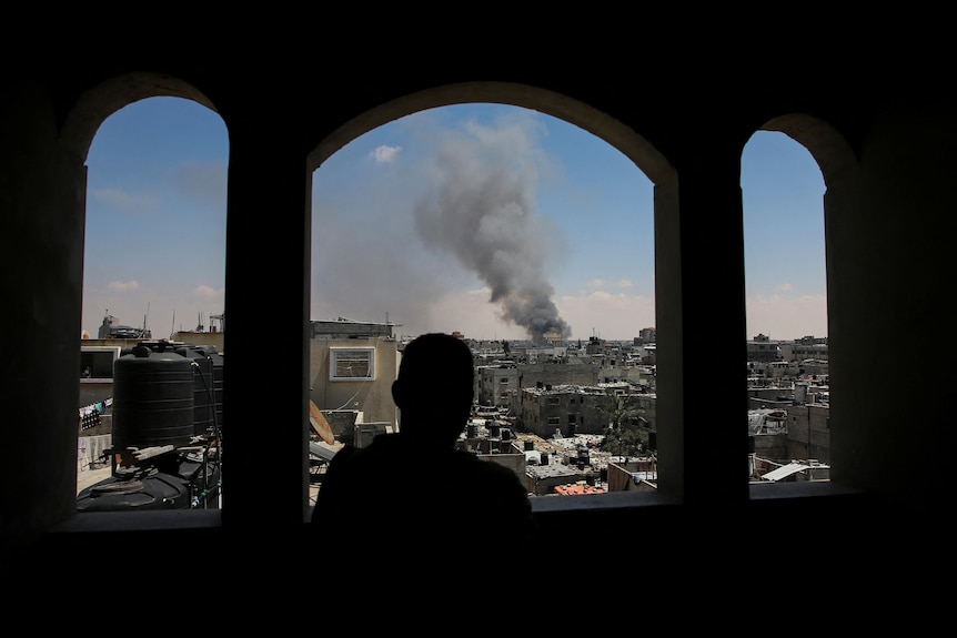 A Palestinian man stands looking through a opening in a building, watching as smoke rises after Israeli strikes.
