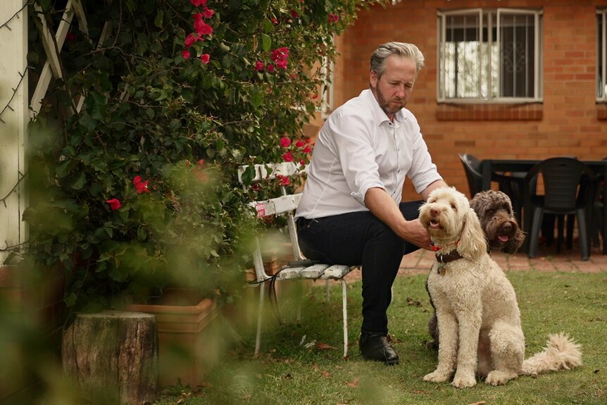 Ben Cannon sitting on a bench infront of his house, patting his two dogs with hedge in foreground