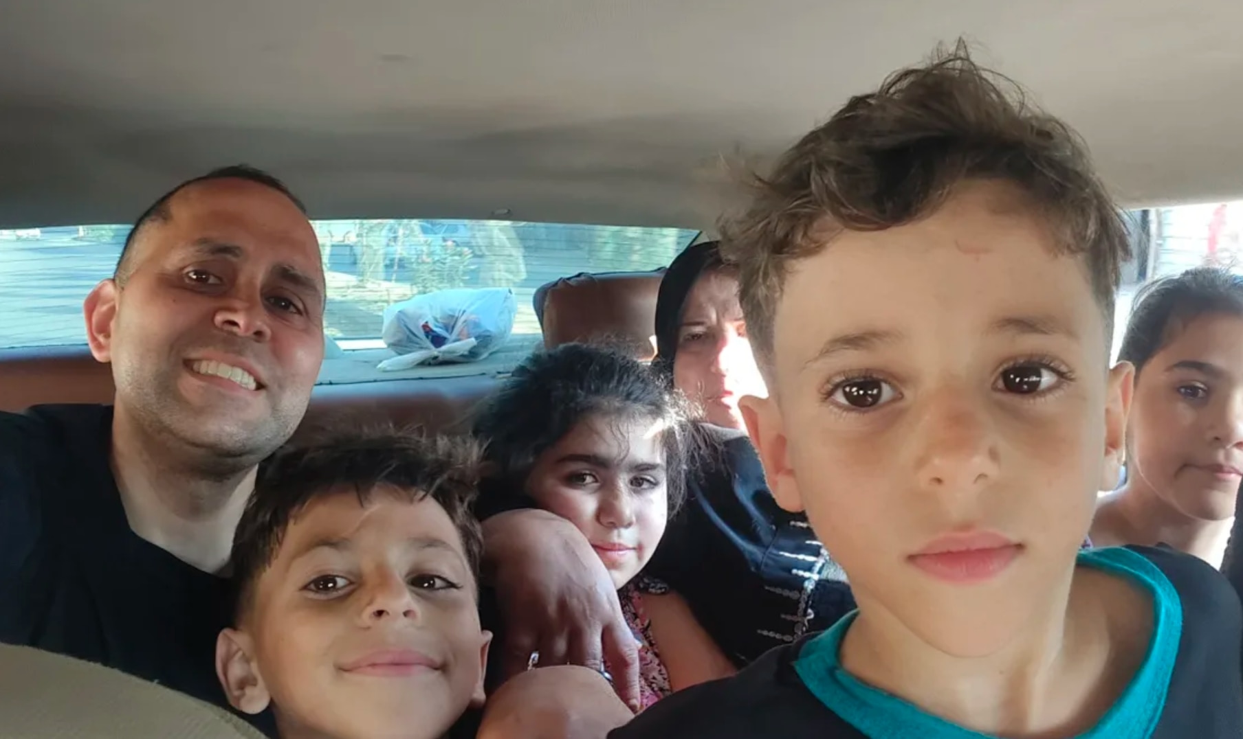 Hani is pictured smiling in the back of a car. There are children around him.
