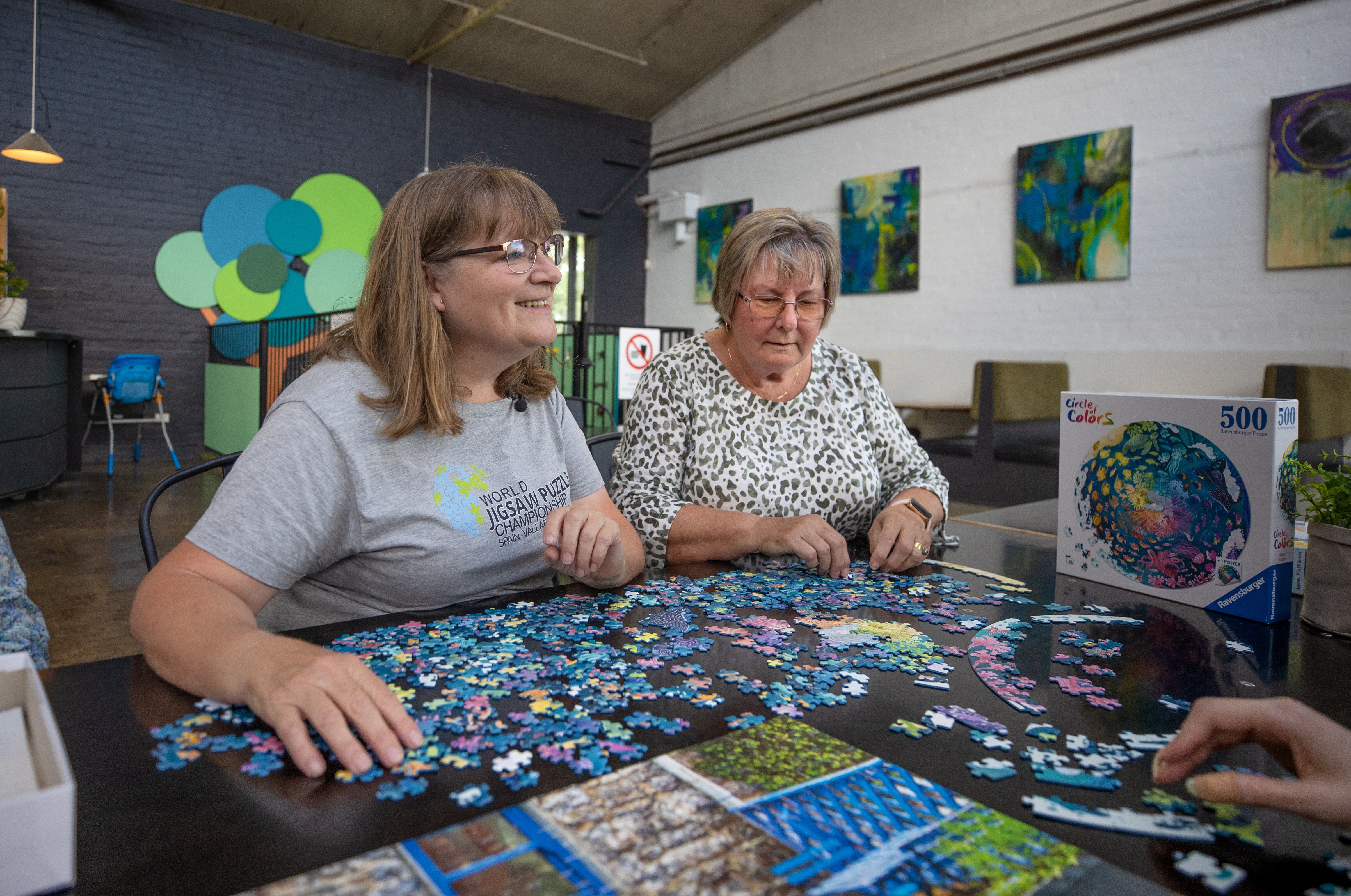 Katrina Coleman and Cathy Kerrison sit next to each other at a table upon which jigsaw puzzle pieces are spread out
