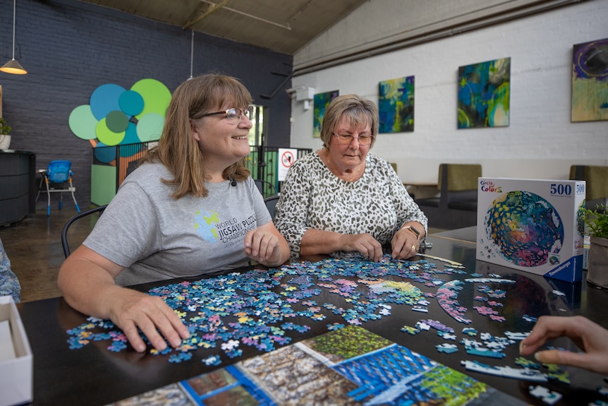 Katrina Coleman and Cathy Kerrison sit next to each other at a table upon which jigsaw puzzle pieces are spread out