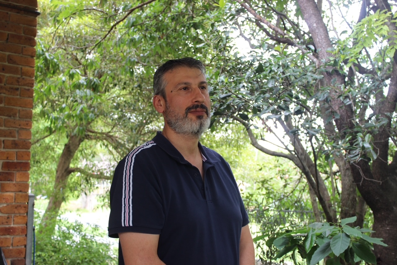 Un hombre barbudo se encuentra en su porche frente a un jardín y posa para una fotografía.