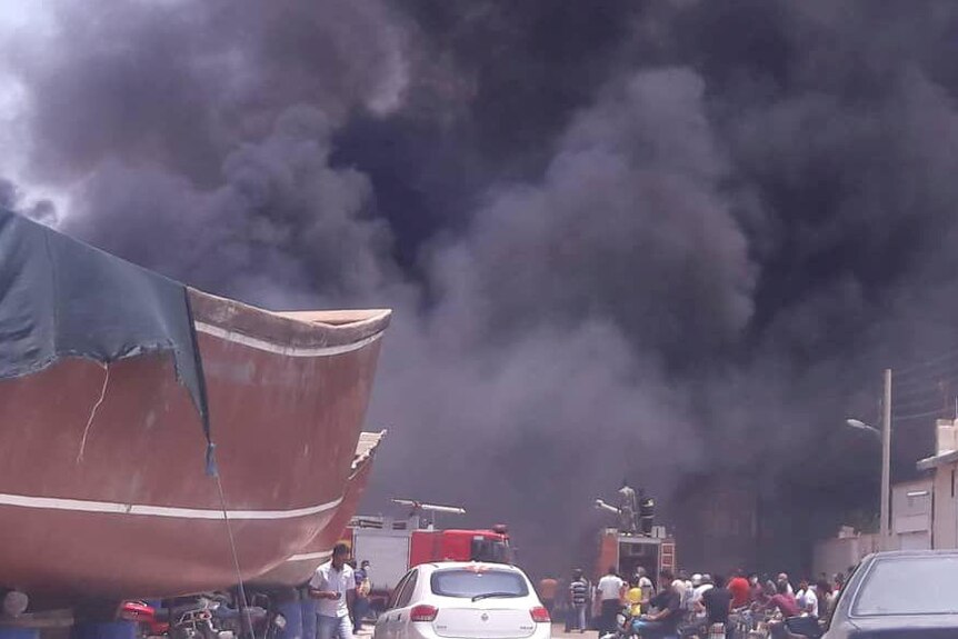 A thick black smoke cloud rises behind parked fire trucks and ship hulls as people huddle around.