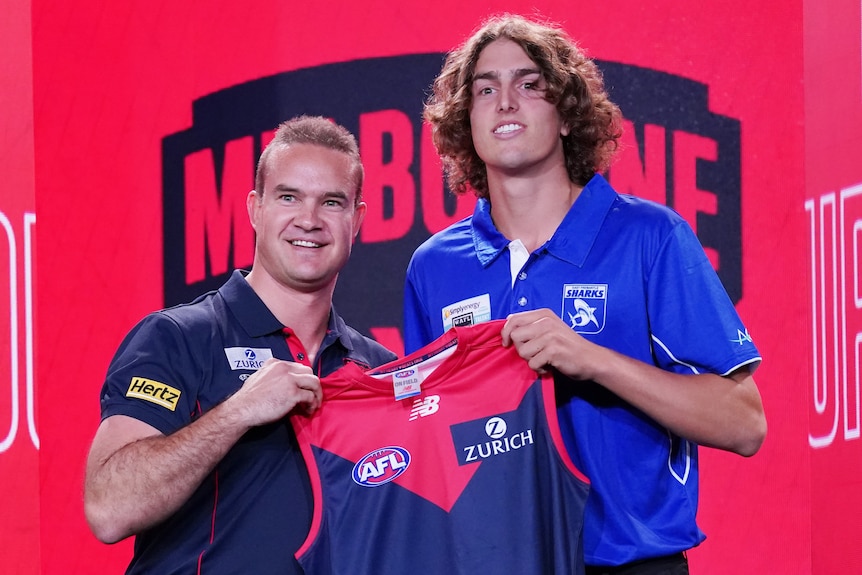 A tall man with long hair stands next to a shorter man while both hold a Melbourne Football Club jumper. 