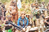 A man wearing a blue jumper and hat sits on a log surrounded by men holding guns.
