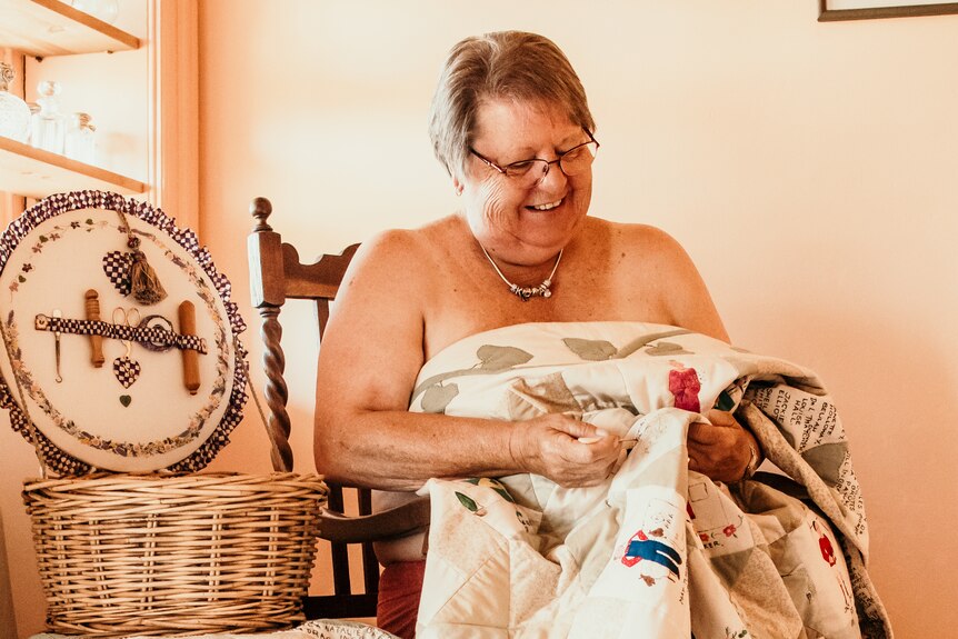 A woman wearing nothing but a quilt, appearing to be knitting. 
