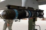 A man in camouflage uniform ducks under the wing of an aircraft carrying a missile about a metre in length