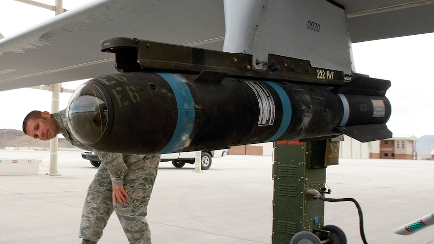 A man in camouflage uniform ducks under the wing of an aircraft carrying a missile about a metre in length