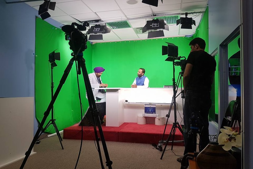 Mintu sits at a desk with another Sikh man in traditional garments in front of a green screen and film equipment.
