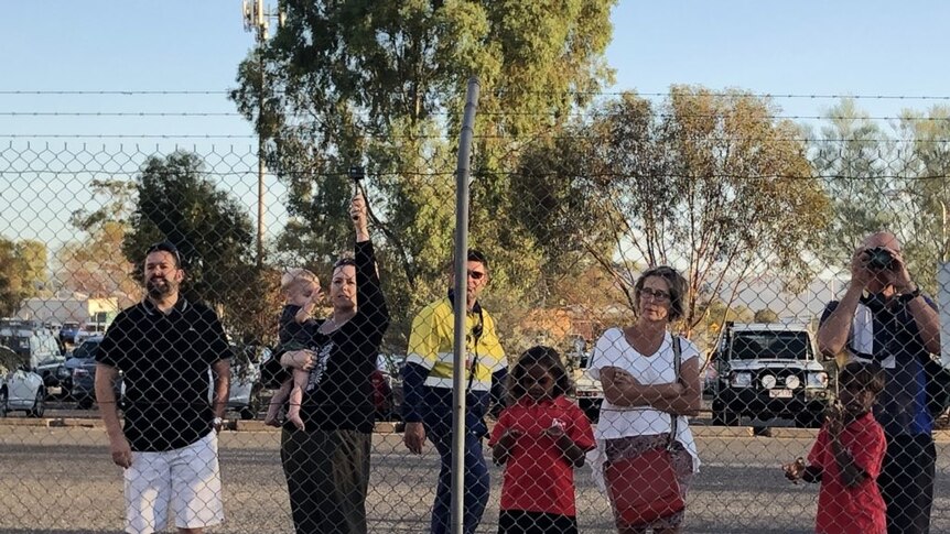 Eight people stand at a fence with barbed wire on top at an Alice Springs car park.