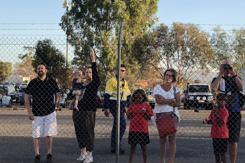 Eight people stand at a fence with barbed wire on top at an Alice Springs car park.