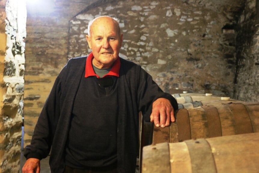 A older man wearing a red shirt under black robes, standing next to some wine barrels.