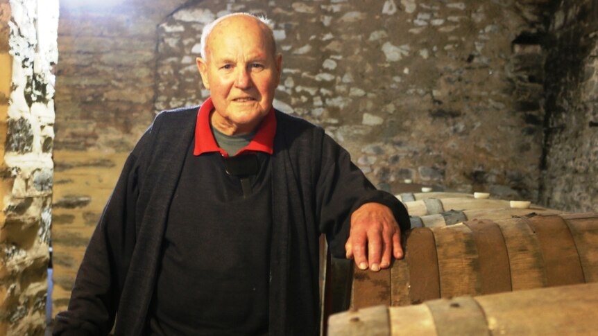 A older man wearing a red shirt under black robes, standing next to some wine barrels.