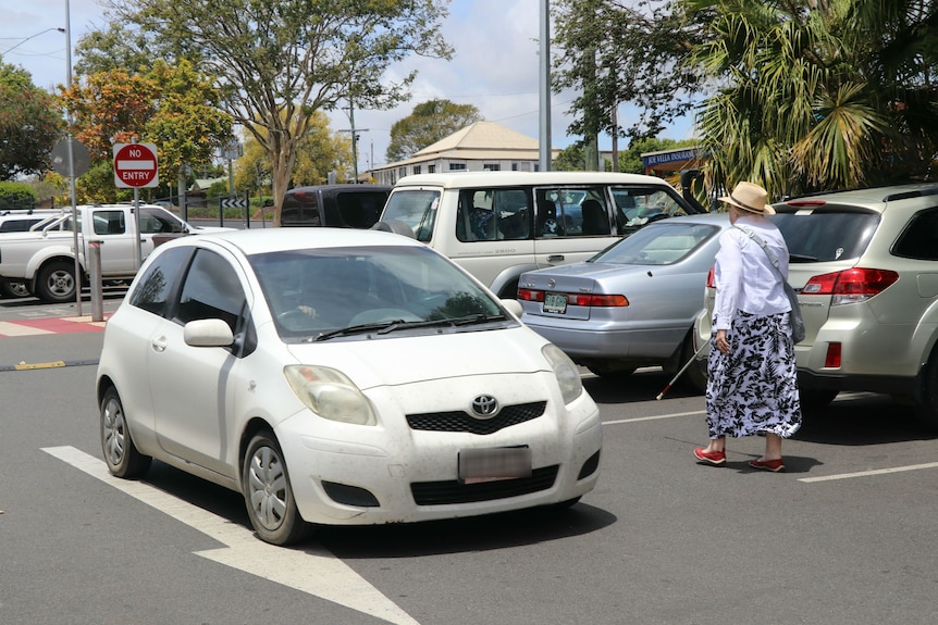 A woman with a white cane walks beside a car and parked cars in a carpark.