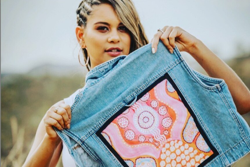 A woman holds a denim vest with a bright print on the back