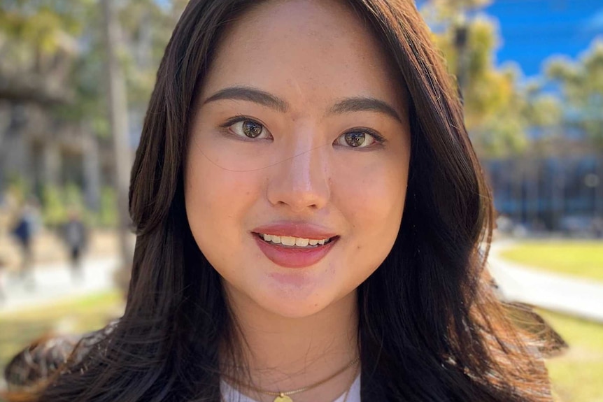 Abbey is standing in a park at the University of Sydney, she is smiling and wearing a black jacket with a white shirt on.