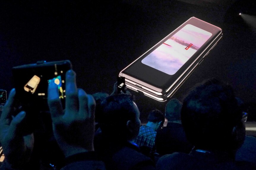 Mobile phones are held up to take a photo of a new model of phone presented on a screen sitting on a black dais